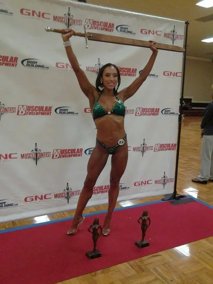Gracelyn on competition stage in green sparkly bikini holding a wooden plank with three trophies, smiling victoriously