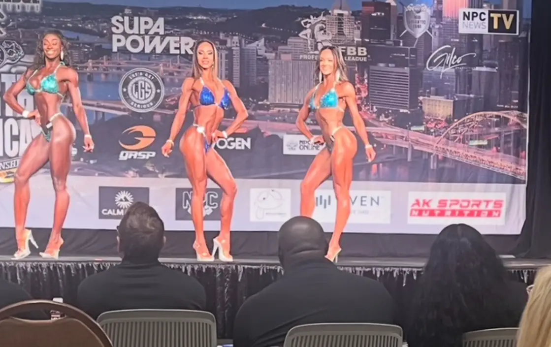 Three bikini competitors on stage under bright show lighting at an NPC event