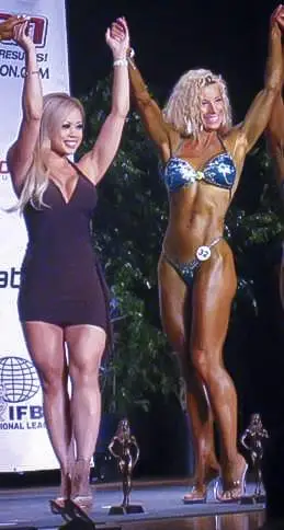 Rebecca winning her first figure competition - on stage in blue sparkly bikini with arms raised in victory, holding hands with the runner-up in a black dress, with IFBB trophies at her feet