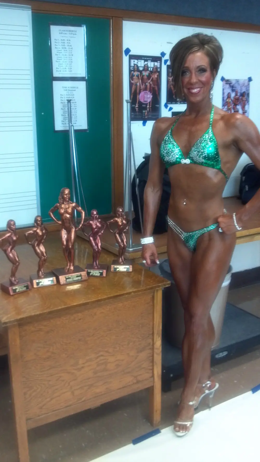 Terra smiling on stage in green sparkly bikini next to her row of competition trophies